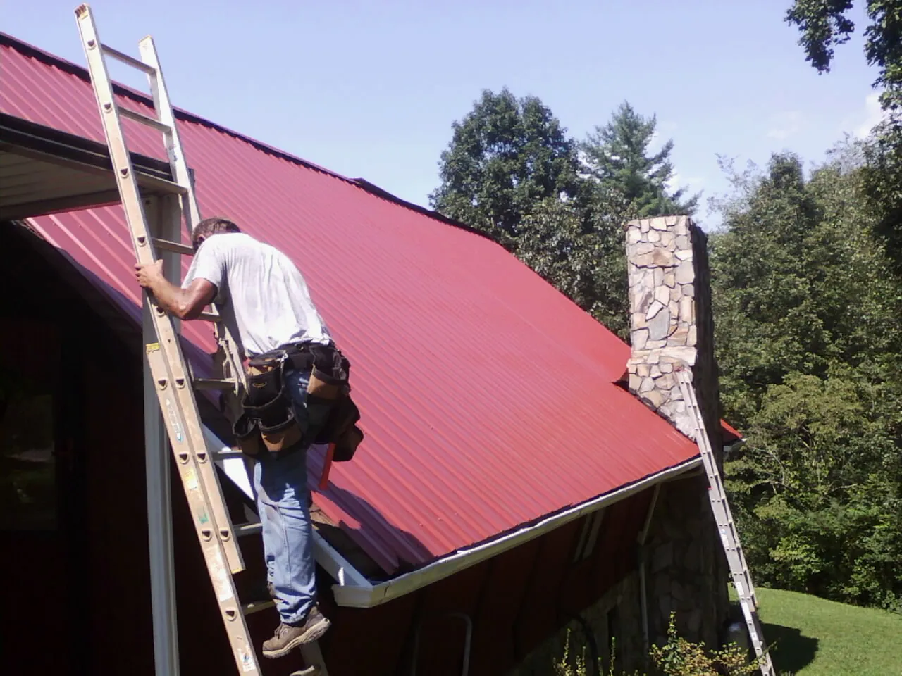 Roofer on a ladder beside a red metal roof and stone chimney — Soffit & Fascia Repair by Roofing Brothers in Glenmont
